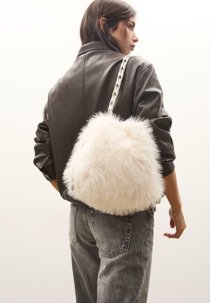 Faux fur taske i off-white, rund form, med fletet rem og lynlåslukning. Den teksturerede overflade tilføjer blødhed og dimension.