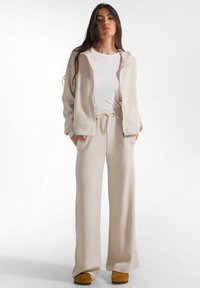 Kvinna med långt mörkt hår som bär beige vida byxor, matchande zip-up hoodie, vit t-shirt och bruna skor, står med händerna i fickorna.