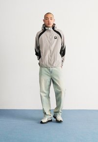 Light gray zip-up jacket with black accents, featuring a hood and logo. Paired with loose, light blue jeans and white sneakers. Plain background.
