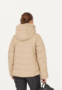 Beige pufferjakke med hætte, der har et quiltet design og en glat tekstur. Modellen holder en sort taske i den ene hånd.