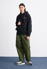 Felpa nera con cappuccio e logo, pantaloni verdi larghi e sneakers grigie con dettagli neri e bianchi. Un outfit casual semplice e funzionale.