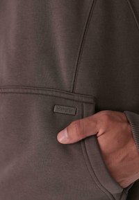 Zbliżenie na dłoń spoczywającą w przedniej kieszeni brązowej bluzy z małą metalową etykietą "Dropsie".