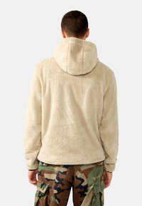 Sudadera de forro polar beige con un bolsillo canguro y capucha con cordón, combinada con pantalones cargo de camuflaje que presentan bolsillos laterales y un corte relajado.