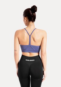 Kvinna sedd bakifrån, bär en lila sport-bh och svarta leggings med "SMILODOX"-märke på midjebandet, håret uppsatt i knut, tatueringar på nacke och arm.