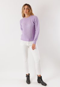 Donna in piedi che indossa un maglione lavorato a maglia lilla, pantaloni bianchi e stivaletti neri contro uno sfondo bianco semplice.
