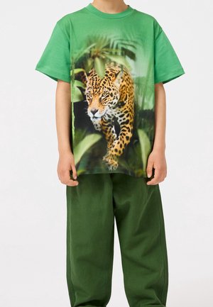 Dítě oblečené v zeleném tričku s realistickým jaguárem procházejícím džunglí vytištěným na přední straně, v kombinaci s tmavě zelenými kalhotami.