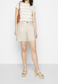 Camiseta blanca a rayas, pantalones cortos beige y sandalias de tacón bloque en colores claros. Detalles notables incluyen un bolso estampado y acentos de botones.