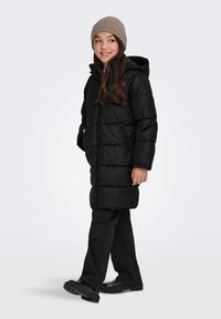 Girl wearing a black patterned puffer coat, black pants, shiny black shoes, and a gray knit beanie, standing and smiling to the side.