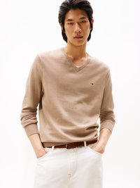Pull à col en V beige à manches longues, en tissu doux. Présente un petit logo sur la poitrine et est associé à un jean blanc et une ceinture marron.