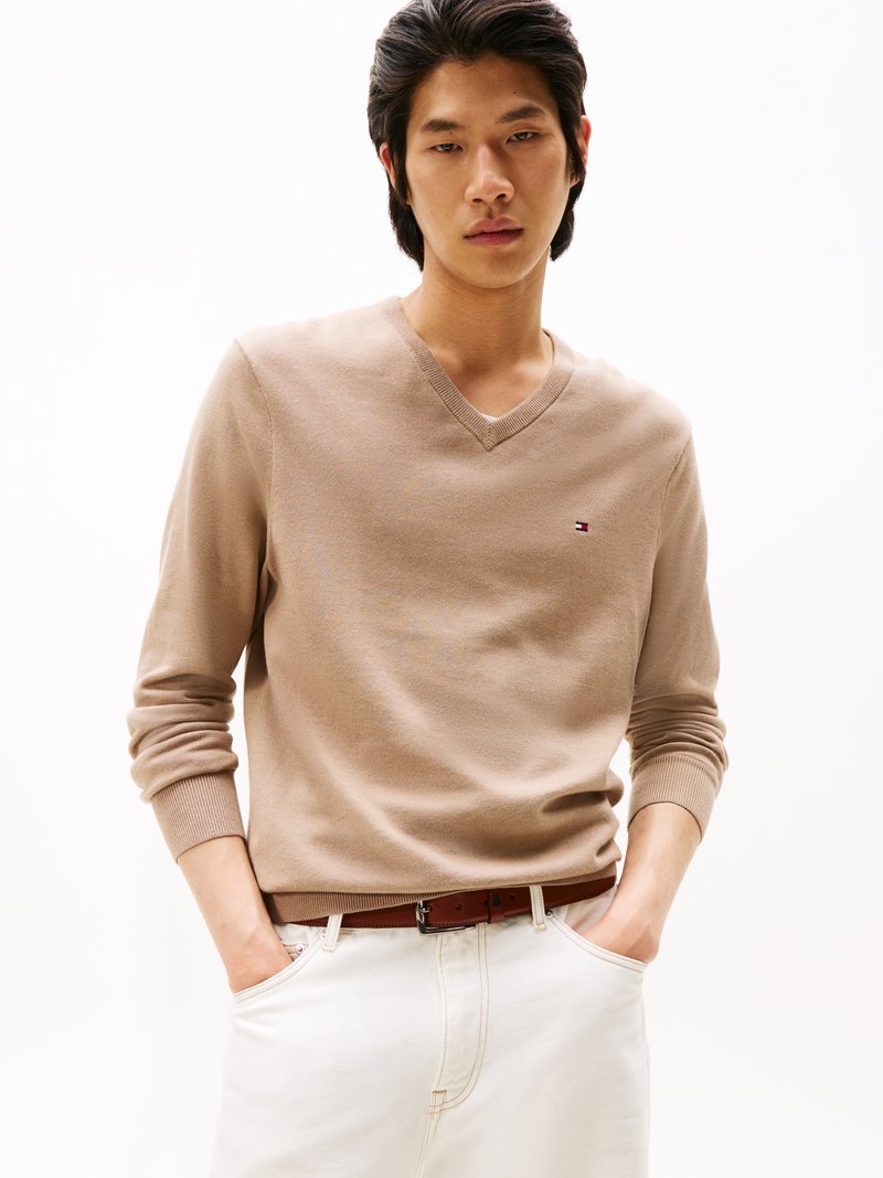 Pull à col en V beige à manches longues, en tissu doux. Présente un petit logo sur la poitrine et est associé à un jean blanc et une ceinture marron.