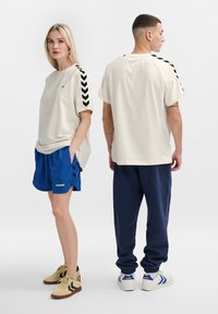 T-shirt beige avec un motif en chevrons noirs sur les manches, associé à un short bleu. Le mannequin porte des baskets beiges, tandis qu'un autre mannequin est vêtu d'un pantalon bleu marine.
