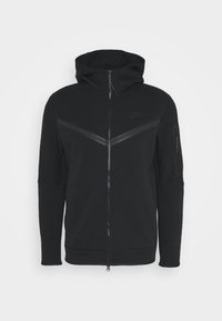 Svart Nike zip-up hoodie med hög krage, utrustad med matta, texturerade chevron-accenter och en dragkedjeförsedd ärmficka. Mjuk tyg.