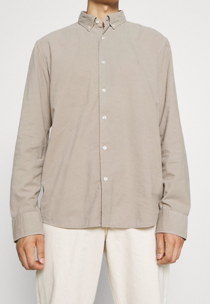 Homme portant une chemise beige à manches longues boutonnée avec un col et un pantalon blanc, debout devant un fond uni.