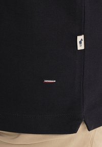 Gros plan sur un vêtement en tissu noir texturé avec une petite bande brodée rouge, blanche et bleue, et une étiquette beige présentant la silhouette sombre d'un cheval.