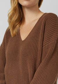 Frau mit langen blonden Haaren, die einen braunen gestrickten V-Ausschnitt-Pullover trägt, dargestellt von der Schulter bis zum oberen Brustbereich.