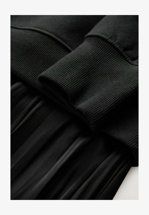 Schwarzer Pullover mit gerippten Bündchen, über einem schwarzen Faltenstoff getragen, der eine glatte Textur und eine einheitliche Farbe aufweist.