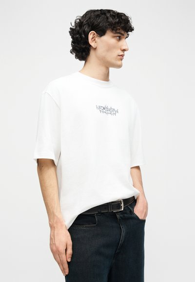 Άνδρας με σγουρά μαλλιά που φοράει λευκό υπερμεγέθες μπλουζάκι με μικρό μαύρο σχέδιο στο στήθος, φορεμένο μέσα σε σκούρα τζιν με μαύρη ζώνη.