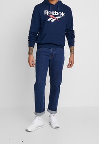 Felpa Reebok blu navy con logo bianco, abbinata a jeans blu scuro e sneakers bianche. Tessuto strutturato, vestibilità regolare, maniche arrotolate.