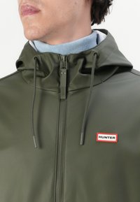 Vihreän oliivinvärinen vettä hylkivä takki, jossa on korkea kaulus, etuvetoketju ja säädettävät kiristysnyörit, sekä näkyvä punainen "HUNTER"-logo.