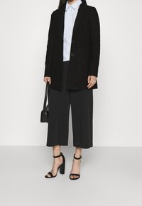 Donna che indossa un blazer nero, una camicia azzurra a maniche lunghe, pantaloni neri larghi e cropped, sandali neri con tacco alto, e tiene in mano una borsa nera.