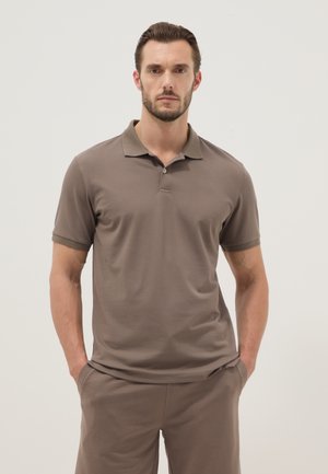 Homme aux cheveux courts et foncés avec barbe, portant un polo marron à manches courtes et un pantalon assorti, debout les mains dans les poches, fond neutre.