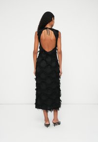 Mujer de pie, vistiendo un vestido midi negro sin mangas con una gran espalda descubierta y patrones circulares texturizados, combinado con mules negros de tacón.
