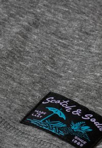 T-shirt din țesătură gri, în prim-plan, cu un etichetă neagră pe care este scris "Scotch & Soda" în culorile purpuriu și turcoaz, reprezentând un design cu umbrelă de plajă.