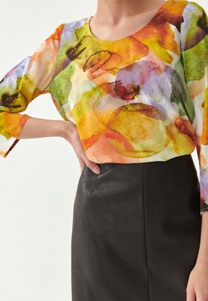 Femme portant un chemisier transparent à motif aquarelle multicolore avec des manches 3/4, rentré dans une jupe en cuir noire taille haute.