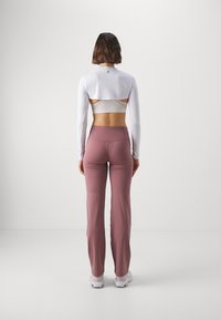 Top bianco a maniche lunghe con dettaglio sulla schiena aperta, abbinato a pantaloni da allenamento a campana rosa chiaro. Tessuto morbido, design aderente e branding minimal.
