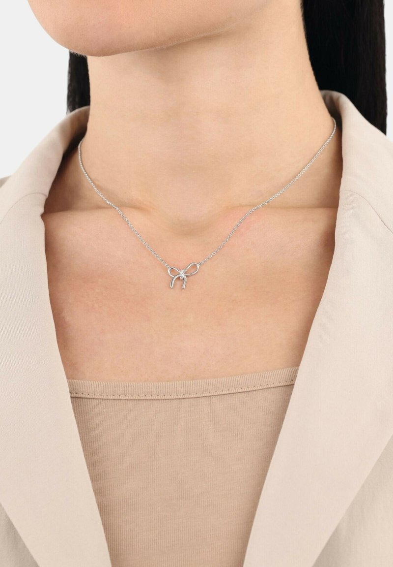 Collana d'argento con un design a fiocco e un piccolo accento, appesa a una delicata catena, indossata su un top dai toni neutri.