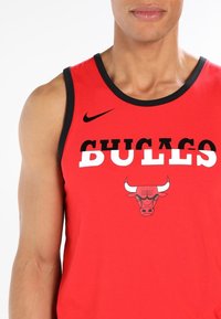 Camiseta deportiva sin mangas roja con ribetes negros, presenta el texto "CHICAGO BULLS" y un logo de toro. Hecha de un tejido ligero. Logo de Nike en el hombro.