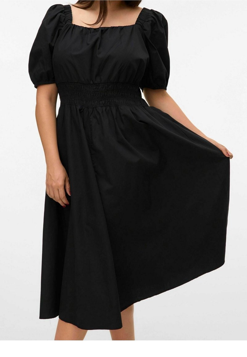 Femme portant une robe midi noire avec des manches bouffantes et une taille smockée, tenant la jupe sur un côté.
