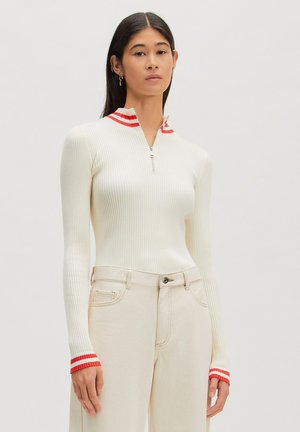 Frau trägt ein cremefarbenes, geripptes Langarmshirt mit rot gestreiftem Kragen und Bündchen, kombiniert mit hoch taillierten cremefarbenen Hosen.