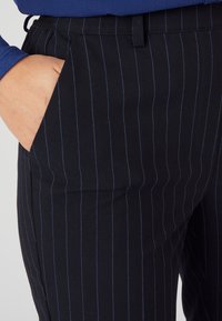 Navy pinstripe broek gemaakt van een gladde stof, met een zijzak en contrasterende blauwe verticale lijnen voor extra detail.