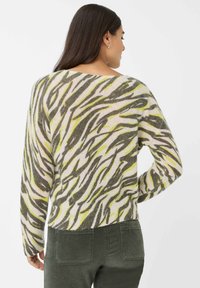 Frau mit langen dunklen Haaren, die einen langärmligen Pullover mit abstraktem Grün-, Grau- und Beigefarbenmuster sowie eine dunkelgrüne Hose trägt, von hinten gezeigt.