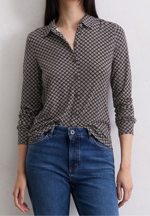 Kvinde iført en sort- og hvidmønstret skjorte med knapper og lange ærmer, stoppet ned i højtaljede blå cowboybukser, stående op ad en væg.