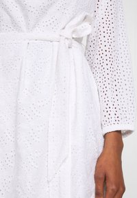 Vestido blanco con un detallado patrón de ojal, mangas largas y un cinturón atado en la cintura. La tela es ligera y texturizada.