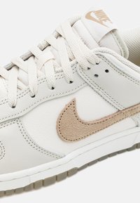Bílá kožená teniska s béžovým semišovým logem Nike, texturovanými krémovými tkaničkami a gumovou podrážkou s průhledným povrchem.