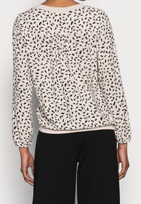 Helles beiges Blusenshirt mit einem schwarzen Blumenmuster, langen Ärmeln, plissierter Rückendetails und einem gerippten Halsauschnitt. Kombiniert mit schwarzen Hosen.