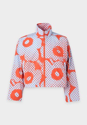 Jeune homme portant un t-shirt blanc, une veste à motif floral rouge et blanc, un jean bleu clair et des baskets blanches, debout avec une main dans la poche.