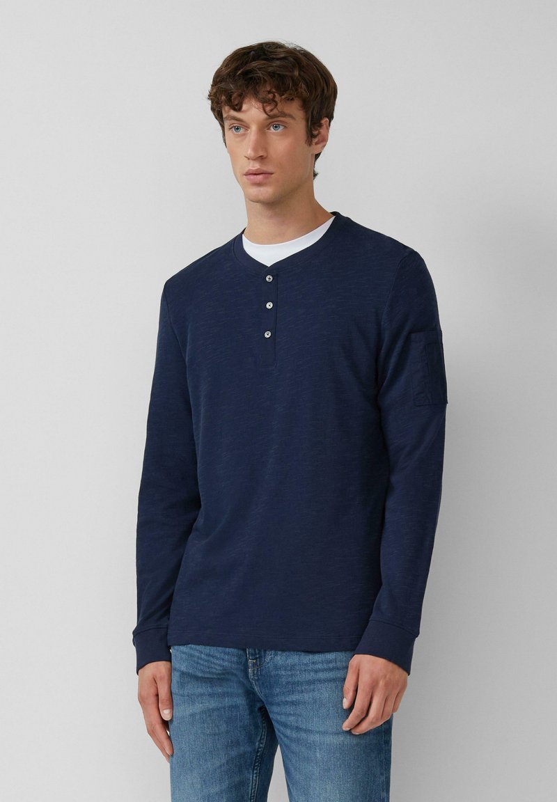 Jonge man in een marineblauwe langarm henley shirt met drie knopen en vervaagde blauwe jeans, staande tegen een effen achtergrond.