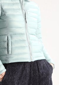 Jasnoniebieska kurtka puchowa z poziomym przeszyciem, całkowitym zamkiem błyskawicznym i kieszeniami po bokach. W zestawie z ciemnymi spodniami w wzory. Gładka tekstura.