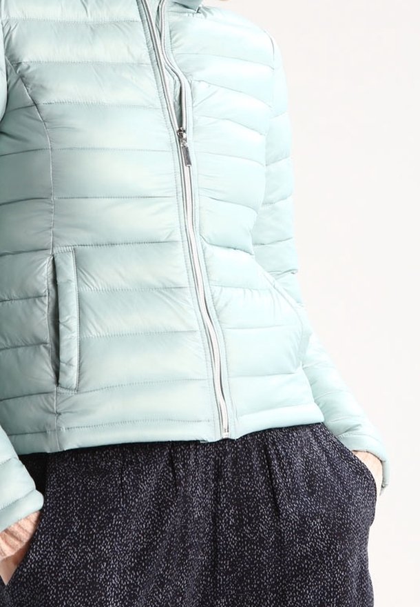 Jasnoniebieska kurtka puchowa z poziomym przeszyciem, całkowitym zamkiem błyskawicznym i kieszeniami po bokach. W zestawie z ciemnymi spodniami w wzory. Gładka tekstura.