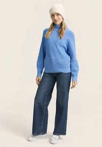 Niebieski prążkowany sweter z wysokim kołnierzem, zestawiony z szerokimi, ciemnymi dżinsami i białymi trampkami. Modelka nosi kremową dzianinową czapkę beanie.