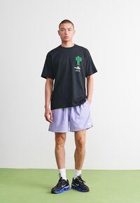 Sort t-shirt med et grønt kaktusgrafik, parret med lilla shorts. Modellen bærer sorte sneakers med blå detaljer og hvide sokker på en grøn overflade.