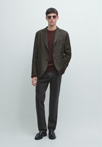 Blazer marrone texturizzato con revers, maglione rosso scuro, pantaloni scuri e mocassini neri. La modella indossa occhiali da sole quadrati, posando davanti a uno sfondo chiaro.