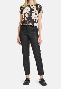 Černá květinová blůza s krátkými rukávy, která má nařasený výstřih. Černé slim-fit kalhoty se zipovými kapsami a lesklé černé boty.