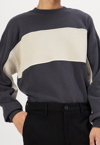 Sweter crewneck w ciemnoszarym kolorze z beżowym, zakrzywionym paskiem. Wykonany z miękkiego materiału, o luźnym kroju i ściągaczach przy rękawach. Połączony z czarnymi spodniami.