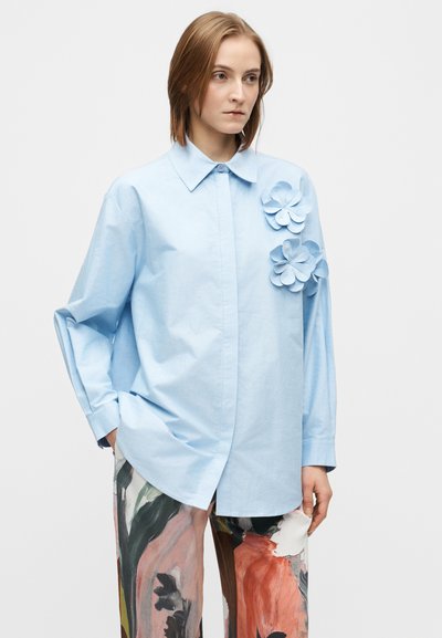 Cămașă supradimensionată de culoare albastru deschis, cu guler cu nasturi și trei accente florale din material pe pieptul stâng. Textură netedă, mâneci lungi.