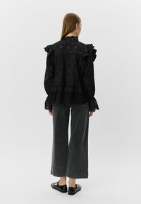 Blusa negra bordada con volantes, detalles recortados y cuello alto, combinada con pantalones grises de pierna ancha y zapatos negros.